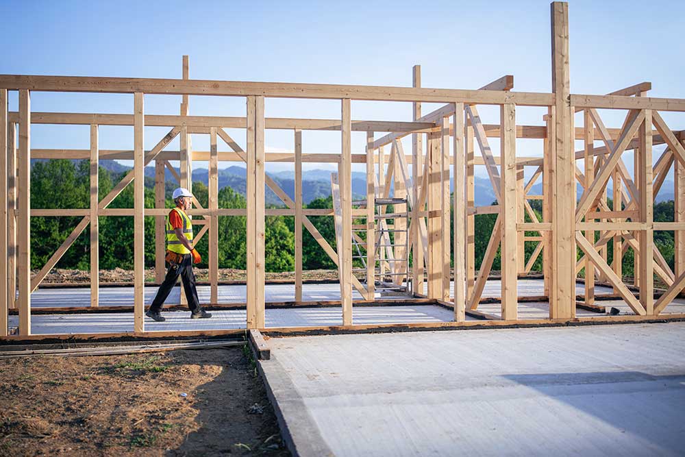 En byggarbetare i gul reflexväst och skyddshjälm går genom träramverket av ett hus under konstruktion